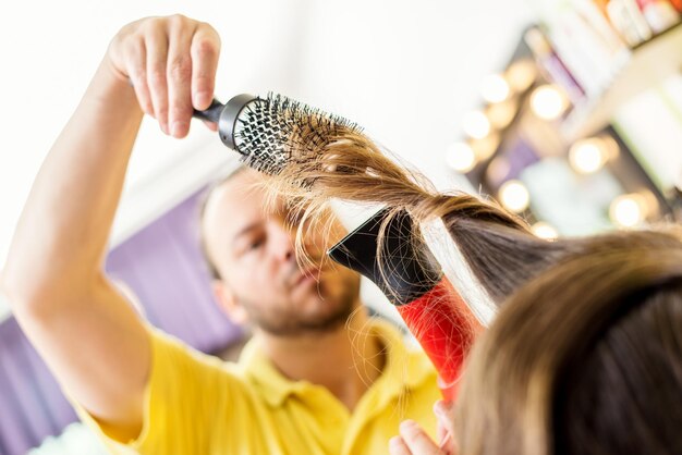 Man hairdresser drying long brown hair of a woman with hair dryer and round brush.