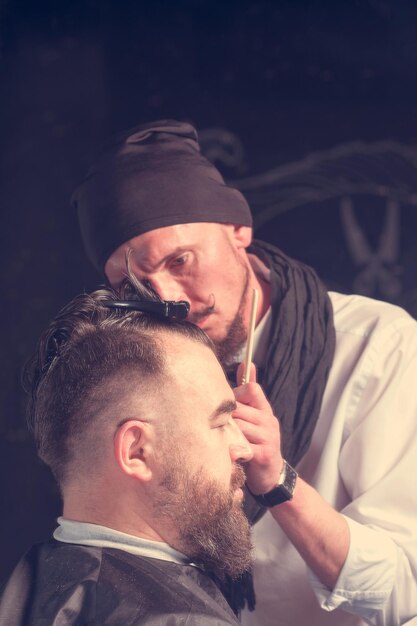 Man hairdresser in a white shirt black hat and scarf doing hairstyle a bearded man with a clip on the head