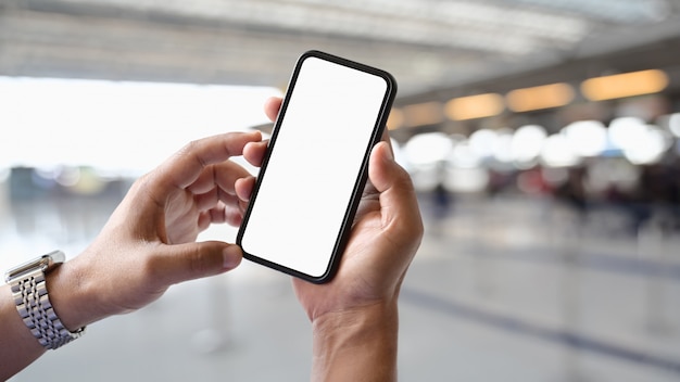 Equipaggi la mano che tiene lo smart phone mobile dello schermo in bianco nel fondo del contatore del terminale di aeroporto