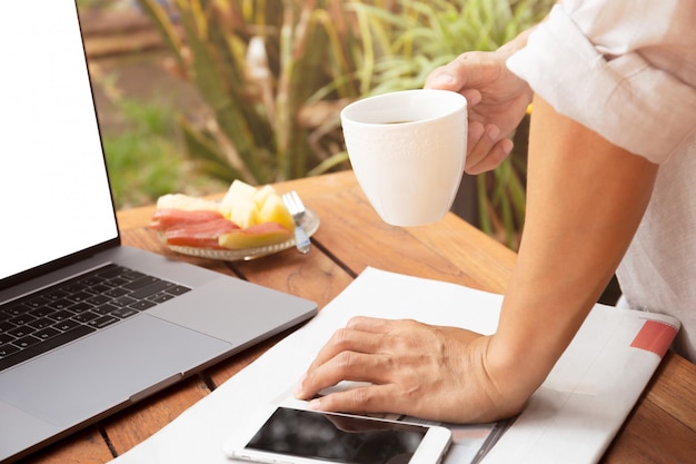 Equipaggi la tazza da caffè della tenuta della mano con il computer portatile ed il giornale, telefono cellulare sul tavolo.