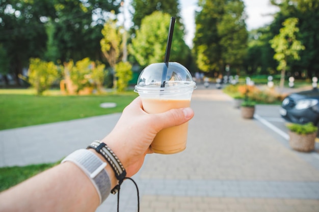 Man hand holding cup with cooling drink walking by summer public park