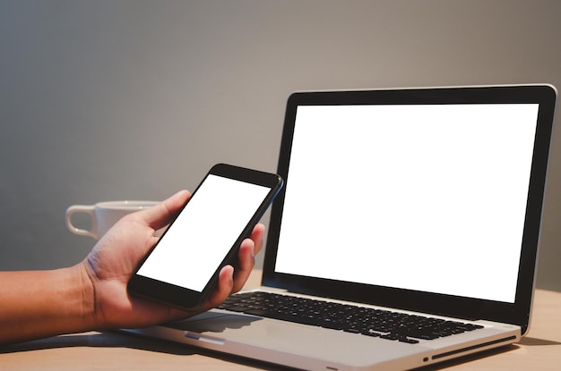 Man hand holding mobile smartphone and computer laptop mock up blank screen on desk