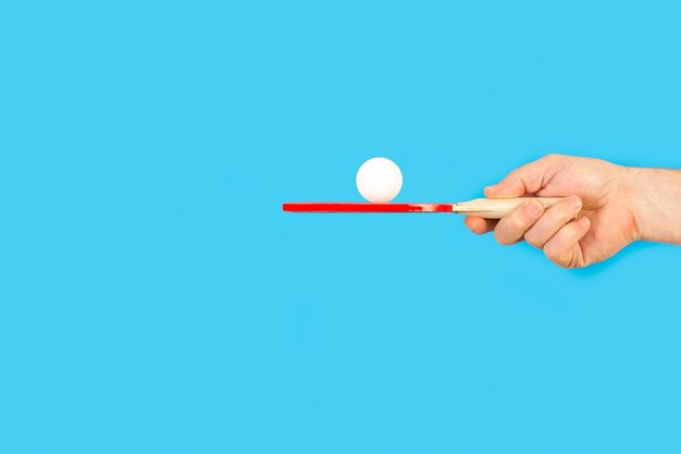 Man hand holding a red ping pong paddle with a white ball on a blue background