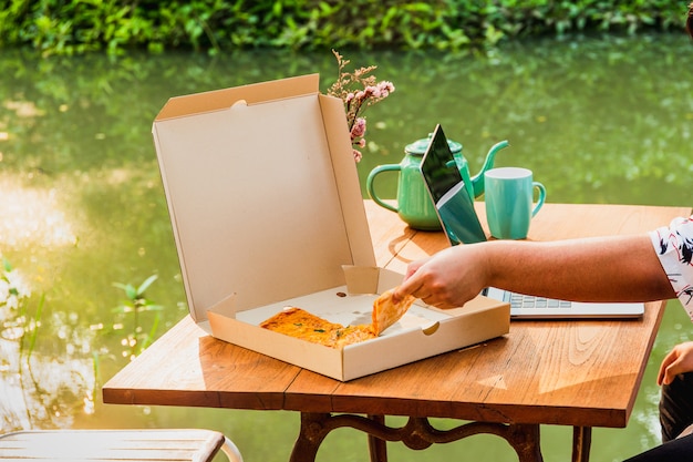 Человек рука держит кусок пиццы и работает на ноутбуке