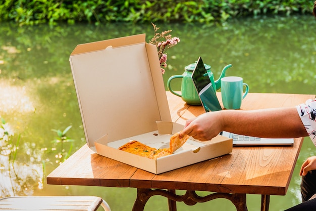 Man hand holding slice of pizza and working on laptop