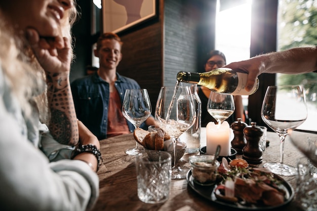 Man hand pouring white wine from the bottle into glasses with\
friends sitting around the table group of young people having food\
and drinks at restaurant