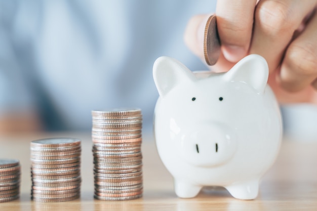 Man hand put coin into White piggy bank with wooden house on wood table saving money buying house