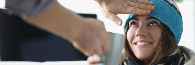 Man hand touching forehead of sick woman in hat and giving cup of tea