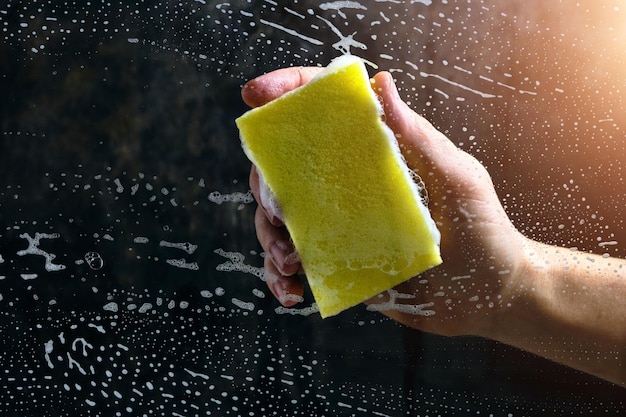 Man hand washes the glass covered with foam, yellow sponge on a dark background.
