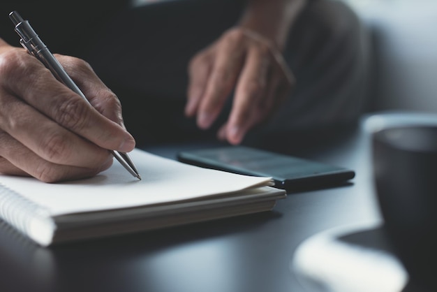 Mano dell'uomo con una penna che scrive su un taccuino di carta e naviga in internet sul telefono cellulare online lavorando sul tavolo in ufficio con sfondo scuro uomo d'affari pianificazione progetto di lavoro sul blocco note da vicino