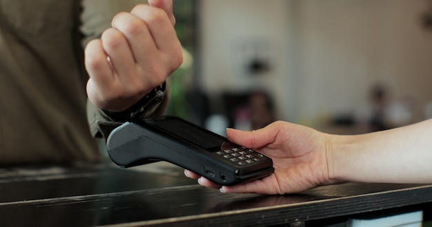 Man hand with smartwatch using terminal for payment in coffee shop, non-cash transaction, side view. Non-cash payment concept. Pos-terminal on table on black background.