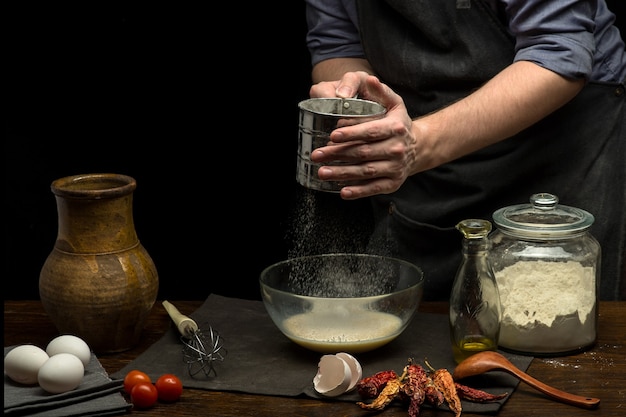 Man hands are pouring flour into glass bowl to prepare dough.