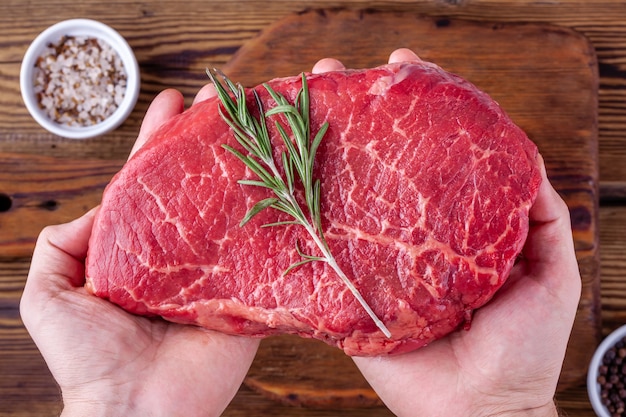 Man Hands Holding a Raw Fresh Beef Steak with Rosemary above a Cups with Herbs