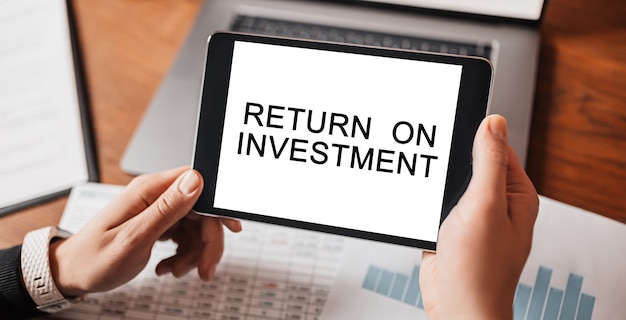 Photo man hands holding tablet with text return on investment at workplace. businessman working at desk with documents