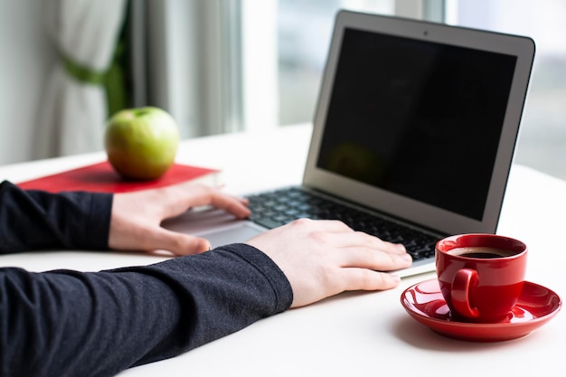 Man hands on laptop keyboard with red notebook, green apple and red cup of coffee on white table