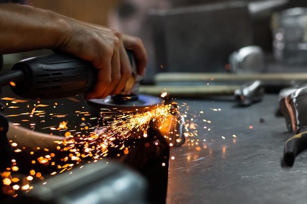 Man mano il trattamento di parti metalliche di hardware in un'officina con smerigliatrice angolare. operaio metallurgico maschio che lucida e finalizza un pezzo di armatura medievale.