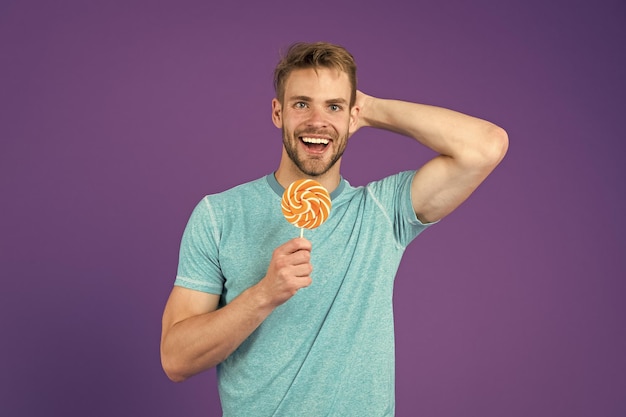 Man handsome macho eat big colorful sweet lollipop Taste of childhood Man bearded adult likes lollipop Sweet tooth concept Sugar harmful for health Guy hold lollipop candy violet background