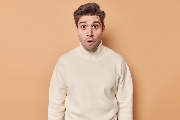 man hapt van verwondering kijkt met ogen vol ongeloof hoort verbluffend schokkend gerucht draagt een casual coltrui geïsoleerd op beige. Omg concept