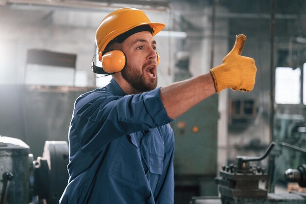 Foto l'uomo con il casco e le cuffie mostra il pollice in alto l'operaio della fabbrica in uniforme blu è all'interno