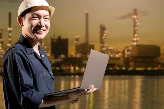 A man in a hard hat stands in front of a gas refinery with a city in the background.