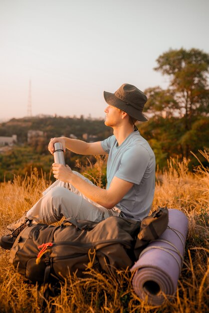 A man in a hat sitting on the hill and opening his thermos