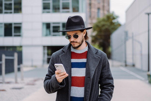 모자와 선글라스를 쓴 남자가 도시에서 야외에서 걸을 때 스마트폰을 사용하여 휴대전화 앱을 사용합니다.