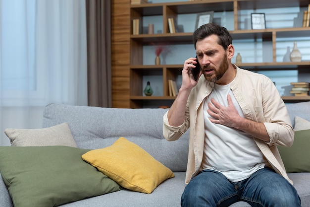 Man having heart attack at home patient calling doctor using phone sitting on couch in living room