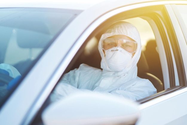 Man in Hazmat suit, protective gloves and goggles driving car during an epidemic in quarantine city.