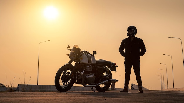 Un uomo con un casco sta accanto a una moto d'epoca su un'autostrada al tramonto.