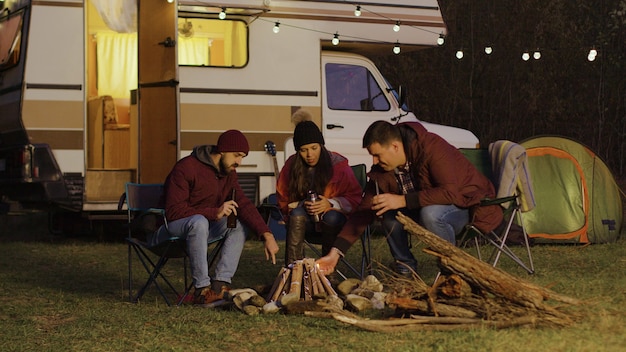 Man helping friend to light the camp fire in front of retro camper. Cold night. Light bulbs in the background.