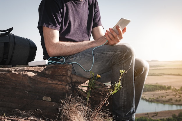 Man on a hike uses smartphone while charging from the power bank on the rock at dawn