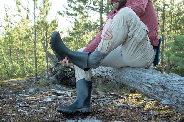 Man in hiking clothes sat on an old log in the woods declines\
rubber boot that shakes dust from it