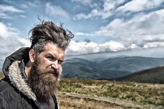Man hipster or guy with beard and moustache on serious face in hat and jacket outdoor on mountain top against cloudy sky on natural background, bearded man