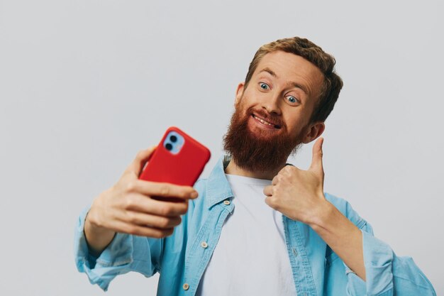 Man hipster met telefoon in de hand blogger glimlach op grijze achtergrond in blauw shirt en wit t-shirt praten aan de telefoon en selfies