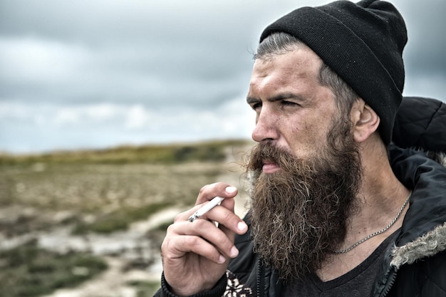 Man hipster of man met baard en snor op ernstig gezicht roken sigaret in hoed en jas buiten op bergtop tegen bewolkte hemel op natuurlijke achtergrond, bebaarde man