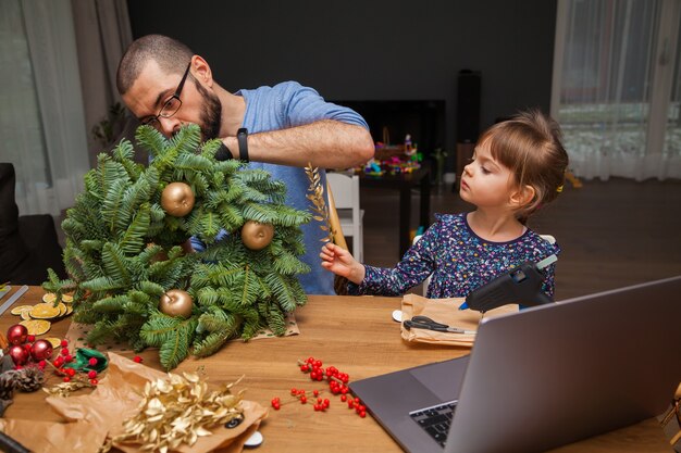 Foto l'uomo e la sua piccola figlia imparano a decorare una ghirlanda di natale guardando la lezione online