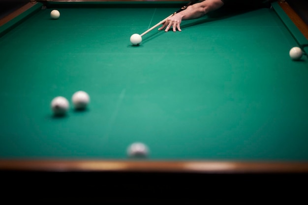 The man hits with a cue a hand with a cue aiming at a billiard\
ball at the table soft focus