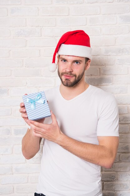 Man hold xmas box with blue ribbon. Macho in santa hat with present on white brick wall. Gift giving, holiday greeting. Boxing day concept. Christmas, new year preparation and celebration.