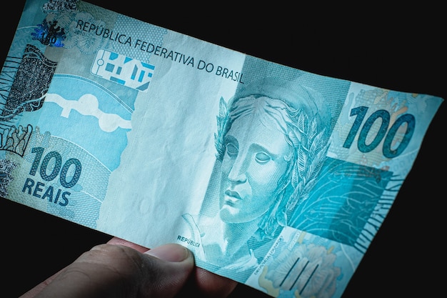 a man holding a 100 bill of brazil money which is the brazilian real on black background