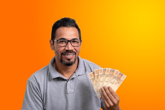 мужчина держит бразильские деньги, оранжевый и желтый фон