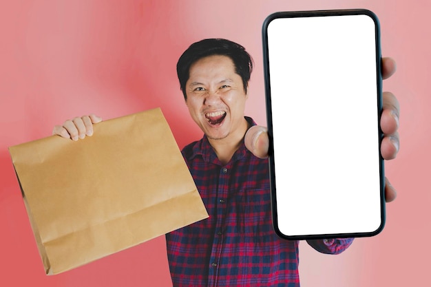 Man holding brown shopping paper bag happy gesture isolated on pink background holding a large smartphone with a blank screen