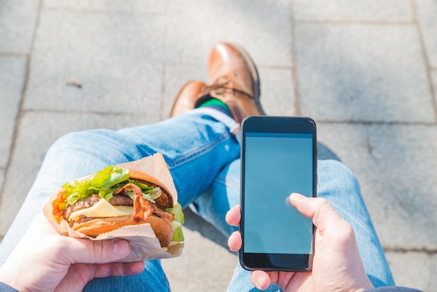 Foto uomo che tiene hamburger e schermo bianco del telefono cellulare