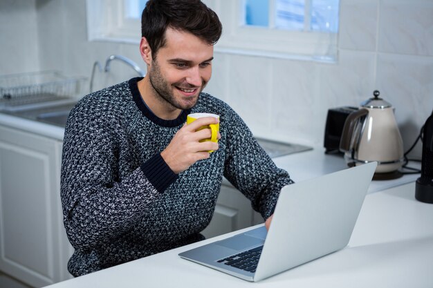 Equipaggi la tazza da caffè della tenuta mentre utilizzano il computer portatile nella cucina