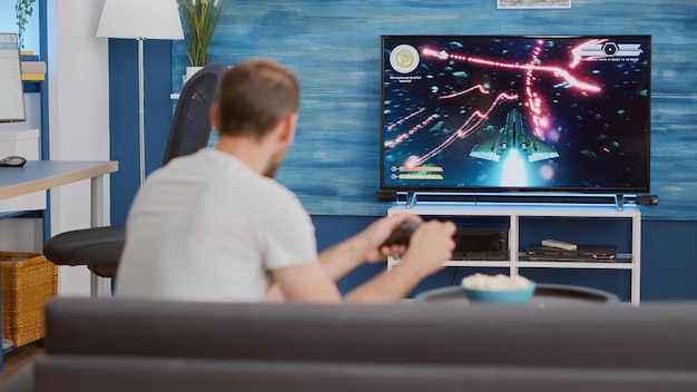 Man holding controller playing action space shooter on console disappointed because losing online game in front of big screen tv. Person sitting on couch unhappy with performance in gaming simulation.