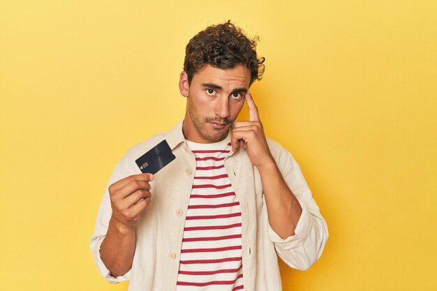 Photo man holding credit card on yellow pointing temple with finger thinking focused on a task