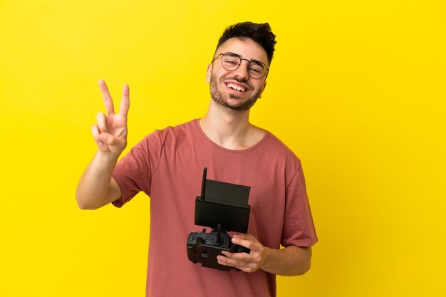 Man holding a drone remote control isolated on yellow background smiling and showing victory sign