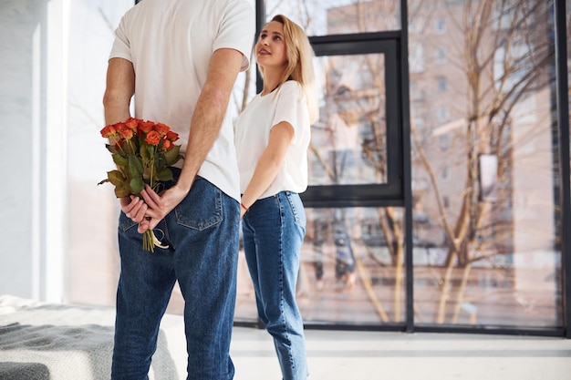 屋内でガールフレンドを驚かせるために背中の後ろで花を手に持っている男