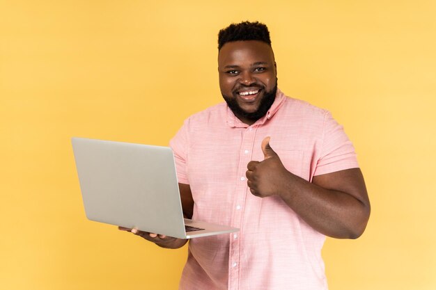 Мужчина держит ноутбук в руке, показывая большой палец вверх, блоггер любит посты, глядя в камеру
