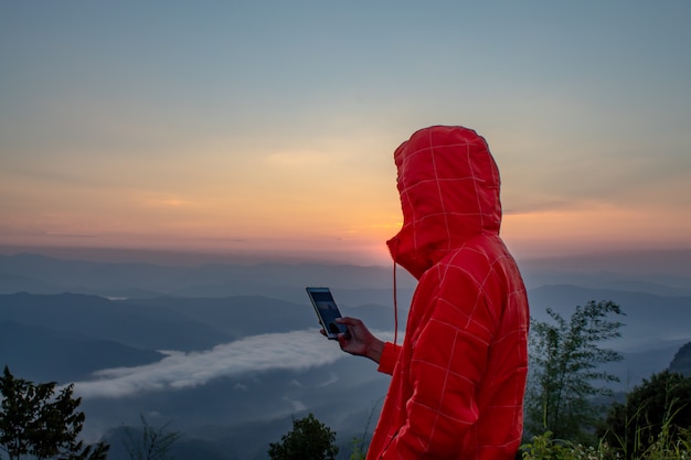 Мужчина держит мобильный телефон на горе с солнцем и туманом.