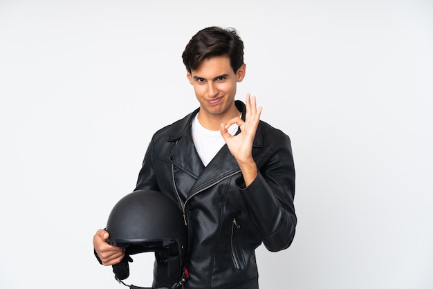 Man holding a motorcycle helmet over isolated white wall showing an ok sign with fingers